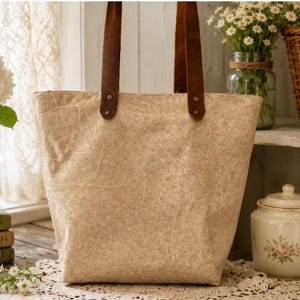 Puede incluir: Un bolso tote de color beige con una superficie texturizada similar al papel y dos asas de cuero marr&oacute;n oscuro sujetas con remaches met&aacute;licos. El bolso tiene una forma rectangular con una base ancha y se mantiene erguido en tono beige.