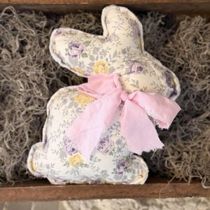 May include: A stuffed bunny rabbit with a floral pattern in shades of purple, yellow, and gray. The bunny has a pink ribbon tied around its neck. The bunny is sitting in a wooden tray filled with dried moss.