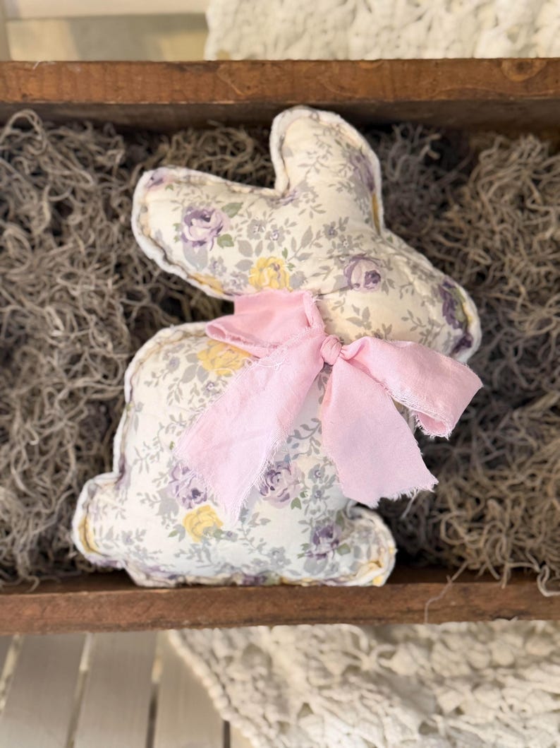 May include: A stuffed bunny rabbit with a floral pattern in shades of purple, yellow, and gray. The bunny has a pink ribbon tied around its neck. The bunny is sitting in a wooden tray filled with dried moss.