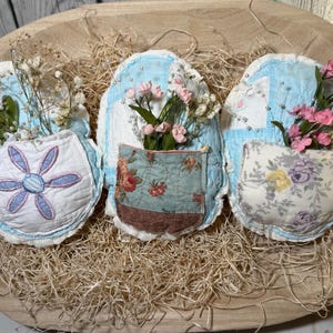 Puede incluir: Tres bolsillos de pared de tela hechos a mano con arreglos florales. Cada bolsillo presenta un diseño único: un patrón de cuadros azules y blancos, un estampado floral y un diseño floral. Los bolsillos están llenos de pequeñas flores rosas y blancas.