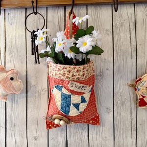 May include: A red fabric wall pocket with a patchwork bird design and the word "TWEET". The pocket is filled with white daisy flowers. A small nest with eggs is below the bird. The pocket is hanging on a wooden wall.