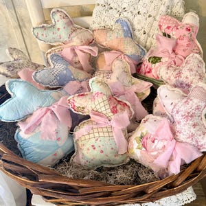 May include: A wicker basket filled with handmade fabric bunny decorations. The bunnies are in various pastel colors and floral patterns, with pink ribbon bows. The basket sits on a surface with dried moss.