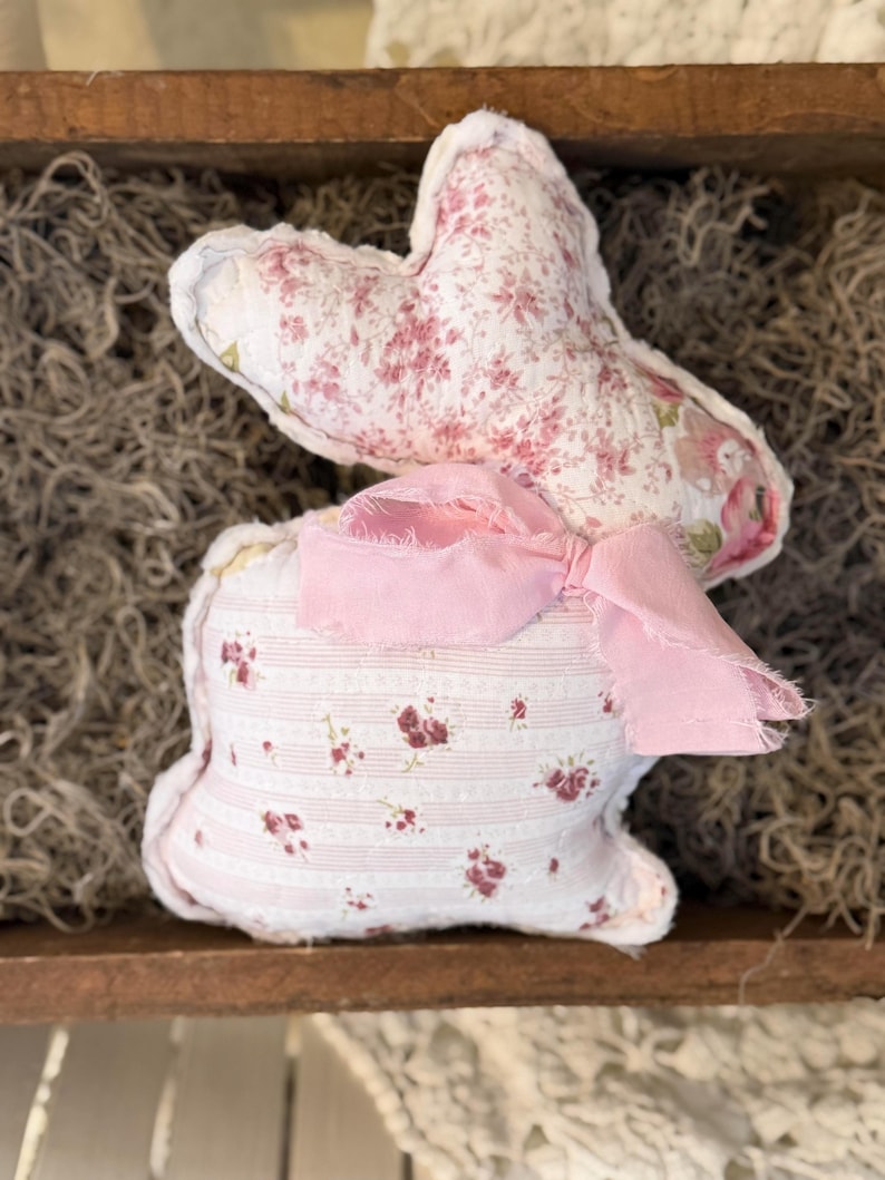 May include: A handmade fabric bunny with a floral pattern in pink and white. The bunny has a pink ribbon bow and is displayed in a wooden tray with dried moss. A charming Easter decoration.