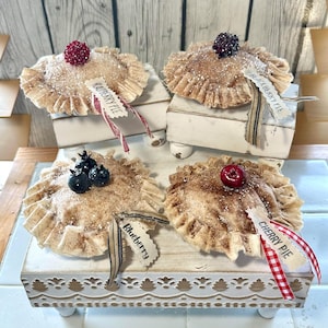 May include: Four decorative miniature pies with ruffled crusts, each topped with a different berry. The pies sit on small white platforms. Tags read "Raspberry Pie", "Blackberry Pie", "Blueberry", and "Cherry Pie".