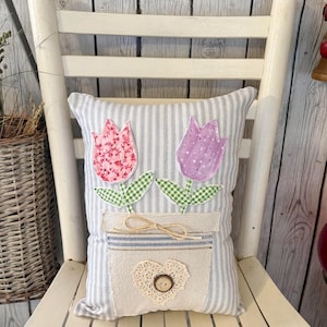 May include: Decorative pillow with a floral design. The pillow features two tulip appliqués, one pink and one purple, with green gingham leaves. The pillow has a striped background and a pocket with a heart-shaped lace detail and a wooden button.