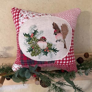 May include: A square patchwork pillow featuring a central circular cross-stitch panel of two birds perched and flying among green holly leaves and red berries. The pillow is constructed from various red and white fabrics, including gingham and floral prints.