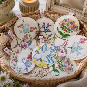 May include: A collection of round, white fabric ornaments featuring colorful embroidered designs of birds and floral clusters. Each circular piece includes a small fabric loop with a metal lobster clasp attached, showcasing detailed stitching in blue, pink, yellow, and green threads.