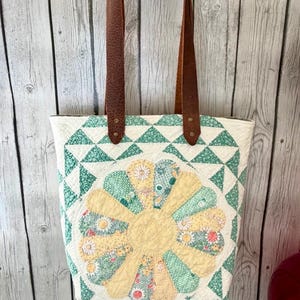Puede incluir: Un bolso tote acolchado con un dise&ntilde;o central de flor amarilla y verde sobre fondo blanco, bordes geom&eacute;tricos de tri&aacute;ngulos verdes y dos correas de cuero marr&oacute;n sujetas con remaches met&aacute;licos en la parte superior.