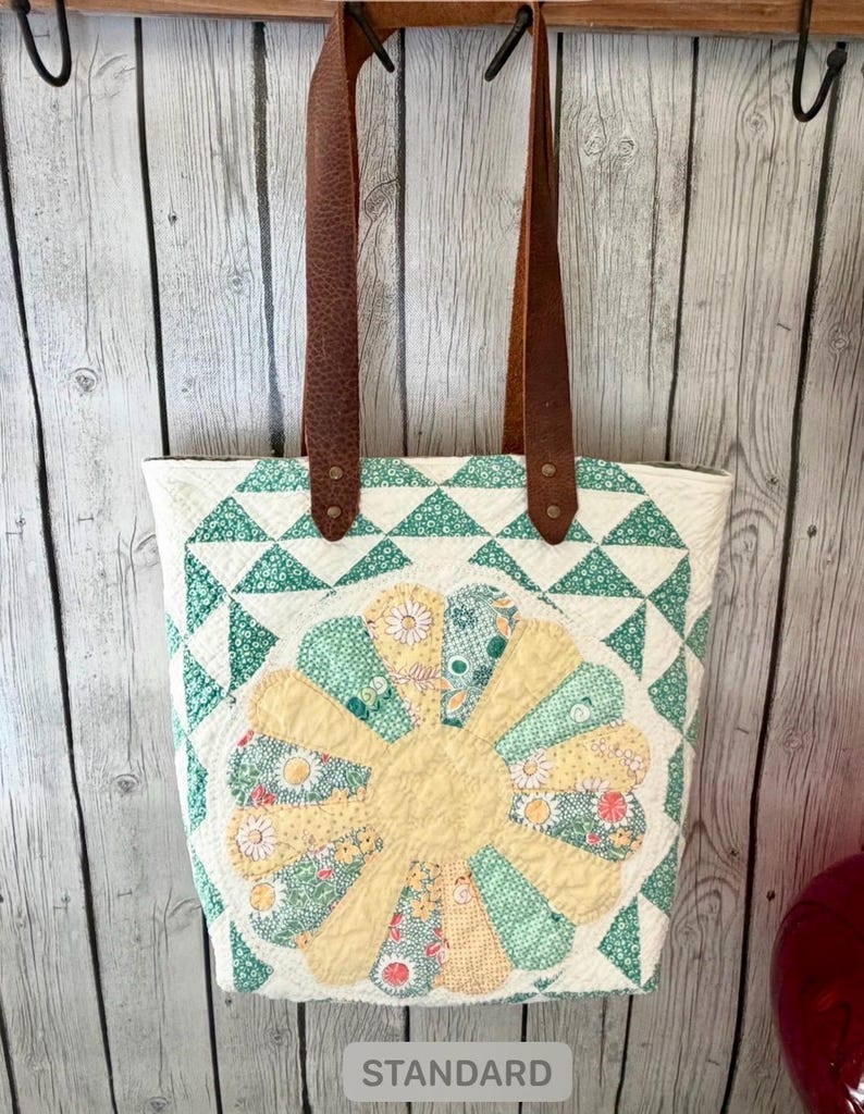Puede incluir: Un bolso tote acolchado con un dise&ntilde;o central de flor amarilla y verde sobre fondo blanco, bordes geom&eacute;tricos de tri&aacute;ngulos verdes y dos correas de cuero marr&oacute;n sujetas con remaches met&aacute;licos en la parte superior.