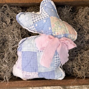 May include: A handmade patchwork bunny in shades of blue and white, with a pink ribbon bow. The bunny is made of fabric squares with floral and geometric patterns. The bunny is a decorative item.