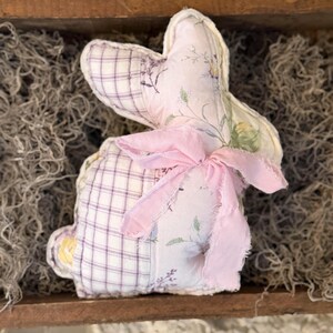 May include: A handmade fabric bunny with a pink bow. The bunny is made of patchwork fabric, with a purple and white plaid pattern on the body and a floral print on the ears. The bunny is sitting in a wooden tray.