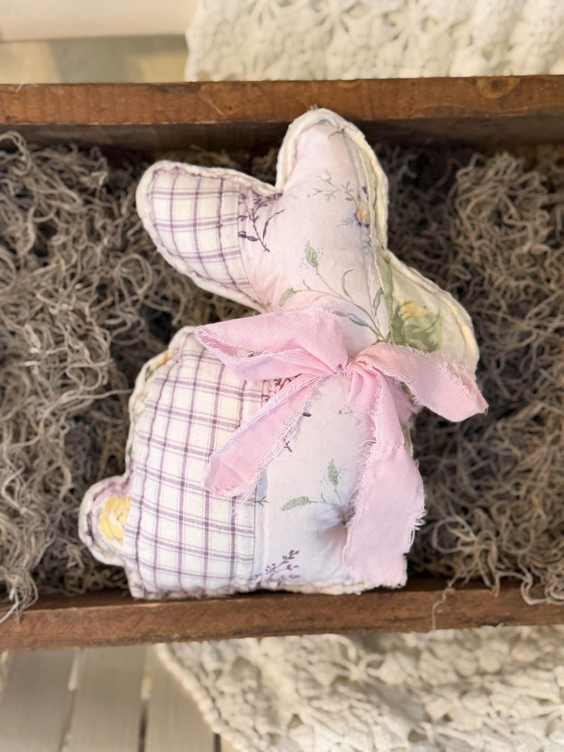 May include: A handmade fabric bunny with a pink bow. The bunny is made of patchwork fabric, with a purple and white plaid pattern on the body and a floral print on the ears. The bunny is sitting in a wooden tray.
