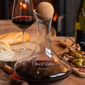 May include: A clear glass wine decanter filled with red wine, with a cork stopper. The decanter has the names "Chloe & Arthur" and the date "12.12.2024" etched on it. A wine glass with red wine, a bottle of wine, and a wooden board with food are in the background.