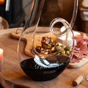Puede incluir: Un decantador de vino de vidrio transparente lleno de vino tinto, con un diseño curvo único. El decantador tiene una inscripción personalizada con nombres y una fecha. El fondo incluye una tabla de madera con aceitunas, nueces y queso.