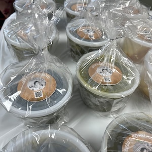 May include: Several plastic containers filled with food, wrapped in clear plastic and tied with a twist tie. Each container has a round sticker with a photo and text. The containers are on a white surface.