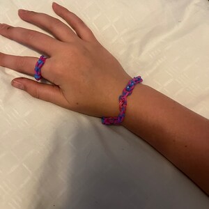 May include: A pink and blue rubber band bracelet and ring. The bracelet is on a person's wrist and the ring is on a finger.