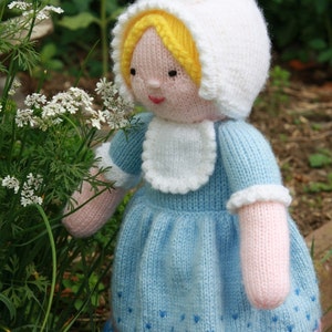 May include: A knitted doll wearing a white bonnet and a blue dress with a white collar and a floral pattern around the hem. The doll has blonde hair and a pink face.