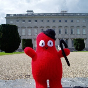 May include: A red knitted toy with a black hat and a black stick in its hand, standing in front of a large, grey building.