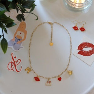 Peut inclure: Un collier en chaîne dorée avec une variété de breloques, dont un cœur rouge, une lèvre rouge, un papillon doré, une enveloppe dorée avec des cœurs rouges et une tasse dorée. Le collier est sur une surface blanche avec un autocollant de lèvre rouge et un autocollant avec la lettre "S" et un cœur.