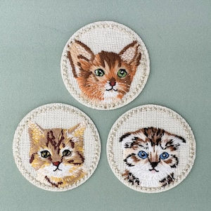 Könnte beinhalten: Drei gestickte Katzenporträts auf runden, cremefarbenen Stoffkreisen mit einer perlenbesetzten Kante. Die Katzen sind alle verschiedener Rassen und haben einzigartige Gesichtsausdrücke.