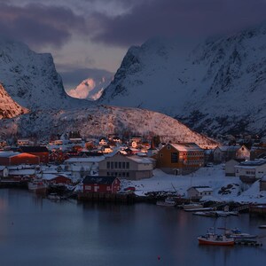Puede incluir: Un pequeño pueblo enclavado entre montañas cubiertas de nieve, con casas rojas y blancas agrupadas alrededor de un puerto. El cielo es de un suave color púrpura y rosa, con un toque de naranja en la distancia. Los barcos están atracados en el puerto, y el agua está tranquila y reflectante.