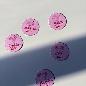 Peut inclure: Ronds autocollants roses avec des illustrations rouges de verres à cocktails et des noms : Maria, Sara, Sophia, Cici et Lucia. Les autocollants sont disposés sur une surface blanche, avec une ombre.