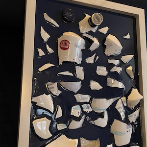 Pode incluir: Uma obra de arte emoldurada que apresenta uma garrafa de vidro quebrada e pedaços de uma garrafa de cerâmica, todos embutidos em uma resina azul escura. As peças da garrafa de cerâmica têm um design branco e azul com o texto "Nordes" e "Gin" visível. A obra de arte é emoldurada em uma moldura branca.