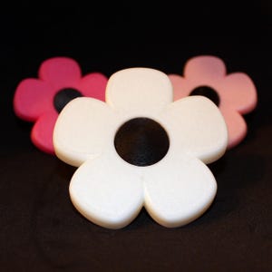 Könnte beinhalten: Drei blütenförmige Objekte in Weiß, Rosa und Hellrosa. Jede Blume hat eine schwarze Mitte. Die Blumen sind auf einer dunklen Oberfläche angeordnet, wobei sich die weiße Blume im Vordergrund befindet.