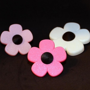 Könnte beinhalten: Drei blütenförmige Verzierungen in Rosa, Weiß und Hellviolett, jede mit einem schwarzen Zentrum. Die Blumen haben eine glitzernde Textur und sind auf einem dunklen Hintergrund angeordnet. Diese dekorativen Elemente könnten für Bastelarbeiten verwendet werden.