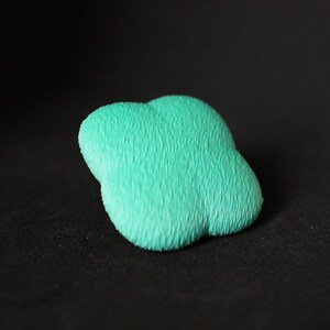 Peut inclure: Gros plan sur un objet en forme de nuage vert clair avec une surface texturée semblable à de la fourrure. L'objet est posé sur un fond sombre, ce qui met en valeur sa forme et sa couleur uniques. Il semble être un objet décoratif.
