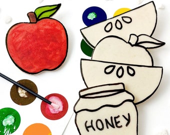 Puede incluir: Un juego de cuatro cortadores de galletas con forma de manzana, tarro de miel y dos rodajas de manzana. Los cortadores de galletas son blancos con contornos negros. El cortador de galletas con forma de manzana es rojo.