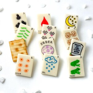 May include: A collection of rectangular, off-white tiles with various hand-drawn illustrations and text. The tiles feature images of insects, boils, frogs, and other symbols. Some tiles have numbers and words like "Seder" and "Frogs".