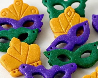 May include: Assortment of Mardi Gras themed masks made of polymer clay. The masks are decorated with purple, green, and gold colors. Some masks are shaped like crowns, while others are shaped like traditional masquerade masks.