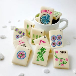 May include: A set of 10 white mahjong tiles with colorful designs. The tiles include numbers 1 through 7, a dragon, a flower, and a joker. The tiles are arranged in a cup.