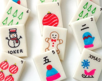 Può includere: Piastrelle rettangolari bianche decorate con motivi natalizi festosi. Le illustrazioni includono un pupazzo di neve, un omino di pan di zenzero, decorazioni natalizie, alberi di Natale e un fiocco di neve con una slitta. Alcune piastrelle hanno numeri e le parole "JOKER" e "XMAS".