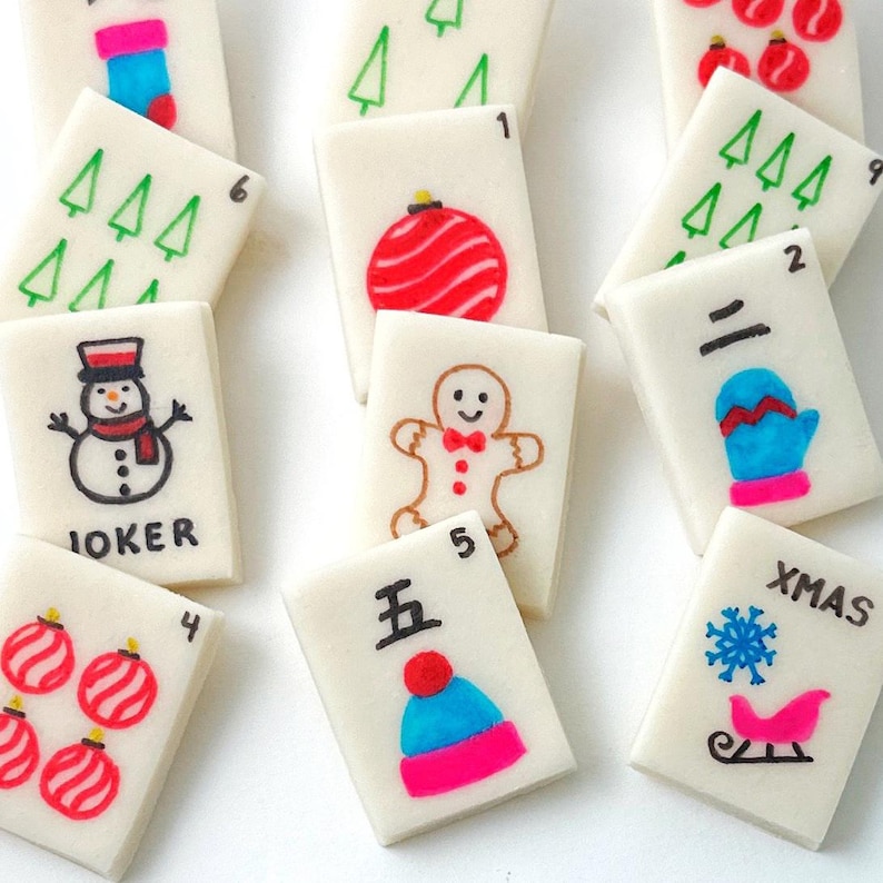 May include: A collection of white rectangular tiles with Christmas-themed illustrations. Designs include a snowman, gingerbread man, ornaments, Christmas trees, and a snowflake with a sleigh. Some tiles have numbers and the word "JOKER" and "XMAS".