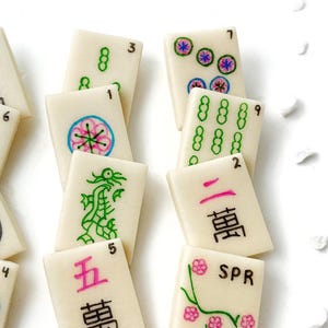 Op de afbeelding: Een set van 10 witte mahjong-tegels met zwarte, groene, roze en blauwe markeringen. De tegels bevatten nummers, Chinese karakters en verschillende symbolen, waaronder een draak, bloemen en cirkels.