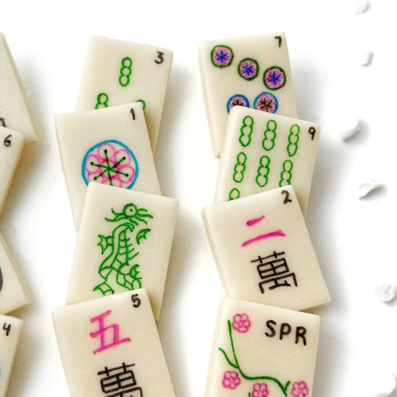 May include: A set of 10 white mahjong tiles with black, green, pink, and blue markings. The tiles feature numbers, Chinese characters, and various symbols, including a dragon, flowers, and circles.