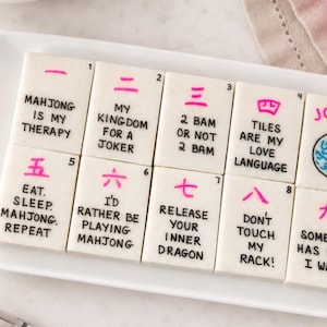 Op de afbeelding: Een set rechthoekige witte mahjongtegels gerangschikt in rijen, elk voorzien van een roze Chinees cijfer, een klein zwart indexnummer en zwarte tekst met zinnen zoals Mahjong Is My Therapy, My Kingdom For A Joker en Don't Touch My Rack.