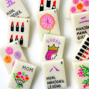 Op de afbeelding: Een verzameling witte Mahjong-tegels met handgetekende illustraties en tekst. De tegels tonen afbeeldingen van lippenstiften, bloemen en het woord "MOM". Sommige tegels hebben nummers en zinnen zoals "MAHJONG LEGEND".