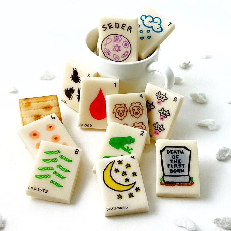 May include: A collection of white, rectangular food items with various illustrations and text, including a red blood drop, a green frog, and the word "SEDER". Some are in a white mug.