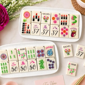 Op de afbeelding: Een verzameling rechthoekige gebroken witte Mahjong-tegels gerangschikt op twee witte rechthoekige dienbladen. De tegels bevatten kleurrijke illustraties waaronder lippenstiften, bloemen, draken, de namen Rachel en Nina en de getallen 37 en 50.