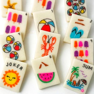 Op de afbeelding: Een verzameling rechthoekige, gebroken witte tegels met kleurrijke illustraties met zomerthema. Ontwerpen omvatten strandballen, ijsjes, een kreeft, een watermeloen en een lachende zon. Sommige tegels hebben nummers en de woorden "JOKER" en "SUM".