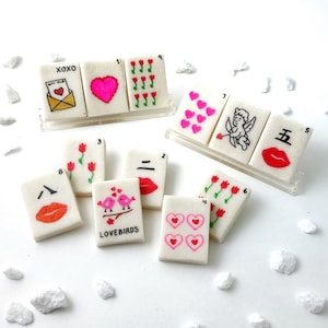 May include: A collection of decorative tiles with various Valentine's Day-themed designs. The tiles feature images of hearts, lips, flowers, and an envelope with the text "XOXO" and "LOVEBIRDS". The tiles are arranged on a white surface with small white rocks.