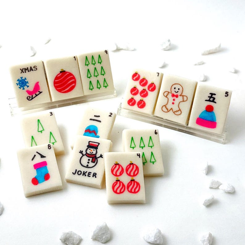 May include: A collection of Christmas-themed Mahjong tiles. The tiles feature festive designs like ornaments, a snowman, a gingerbread man, and a snowflake on a sleigh. Some tiles have numbers and the word "XMAS" on them.