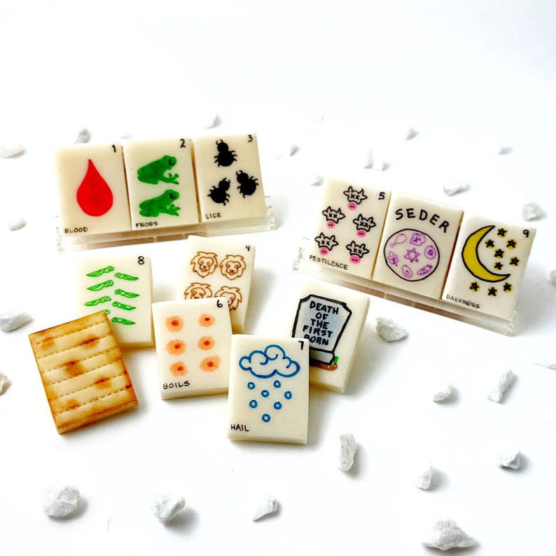 May include: A collection of miniature, cream-colored tiles with various illustrations and text. The tiles depict images such as a drop of blood, frogs, lice, boils, hail, and a crescent moon with stars. The word "Seder" is also visible.