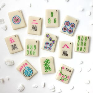 May include: A set of ten white mahjong tiles with various symbols and numbers. The tiles are arranged in a grid pattern on a white surface. The tiles feature a variety of colors, including pink, green, blue, and black.