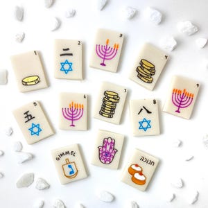 Op de afbeelding: Een verzameling off-white rechthoekige tegels met verschillende illustraties en cijfers. Ontwerpen omvatten een menora, munten, een Davidster en een dreidel. Sommige tegels hebben Hebreeuwse en Chinese tekst. Witte stenen omringen de tegels.