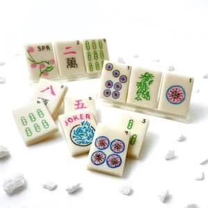 May include: A set of 10 miniature mahjong tiles with various symbols and numbers, including a joker tile. The tiles are white with green, pink, and blue details. The tiles are arranged in a semi-circle on a white surface.