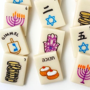 Op de afbeelding: Ivoor-kleurige tegels met diverse handgetekende illustraties en cijfers. Ontwerpen omvatten een menora, een dreidel, munten, een Hamsa-hand en Hebreeuwse en Japanse karakters. Een unieke set voor spellen of display.